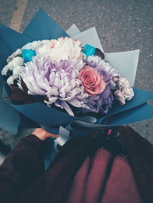 A bouquet of fresh flowers held by a person wearing a dark jacket, featuring a light pink rose, large white and purple chrysanthemums, smaller white blossoms, and bright blue accents, all wrapped in d