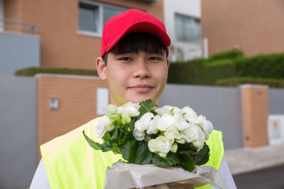 A young delivery person wearing a red cap and a high-visibility yellow vest is standing outdoors in front of modern residential buildings. They are holding a bouquet of fresh white flowers, which incl