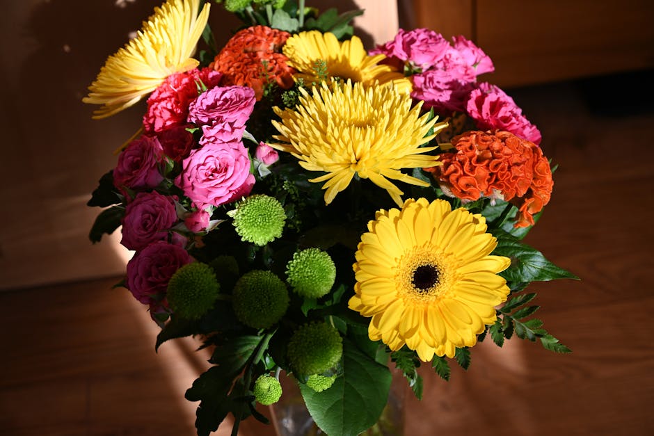 A vibrant floral arrangement featuring large yellow gerbera daisies, pink roses, orange carnations, green pom-pom chrysanthemums, and yellow chrysanthemums, all arranged in a glass vase with lush gree